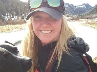 A smiling person with long hair and sunglasses poses with a dog in a scenic outdoor setting.