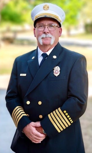 A man in a formal fire chief uniform with a hat and a mustache, standing outdoors with a confident demeanor.