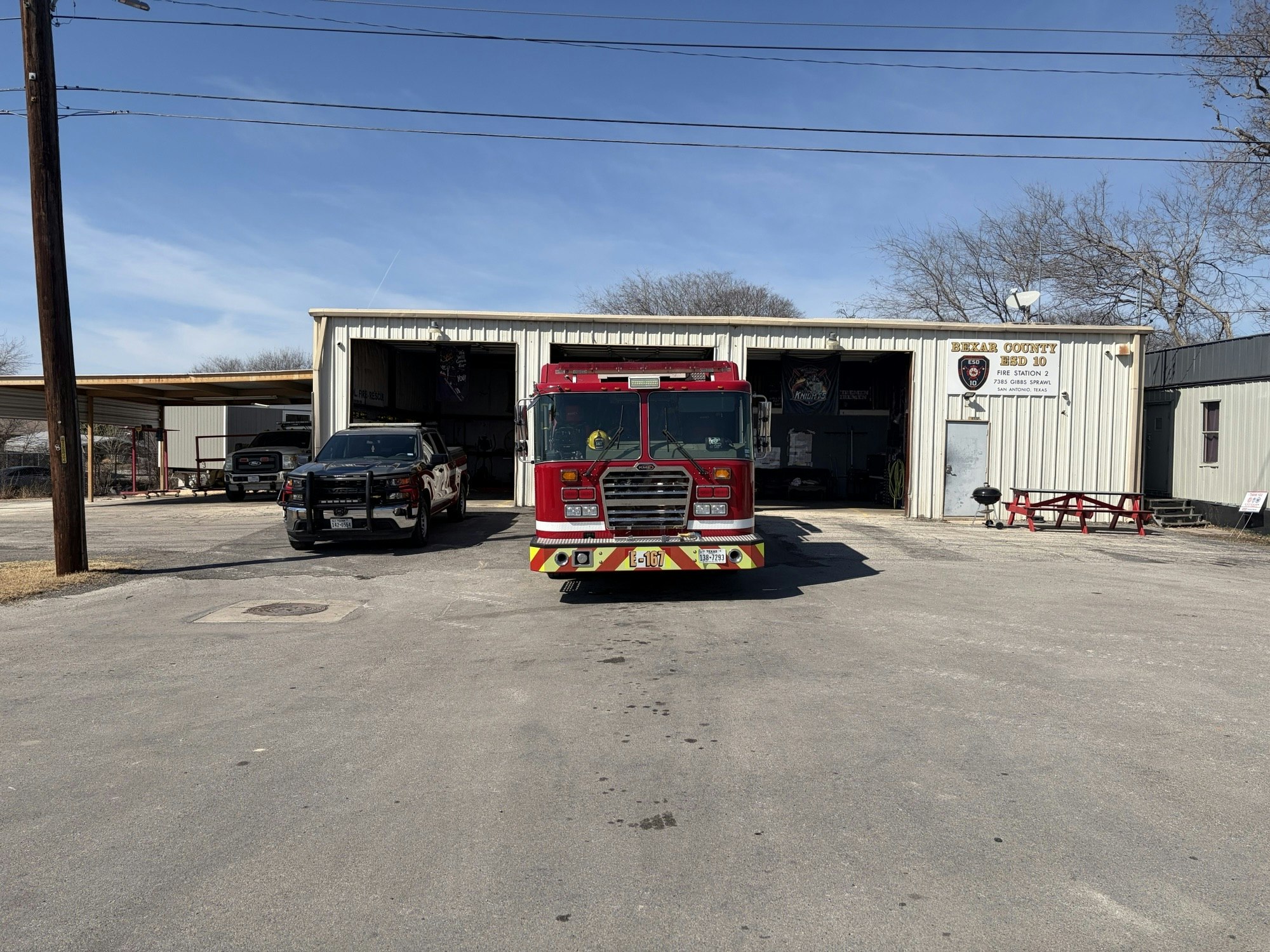 The image shows a fire truck in front of a fire station, with a police vehicle parked nearby and a clear blue sky.