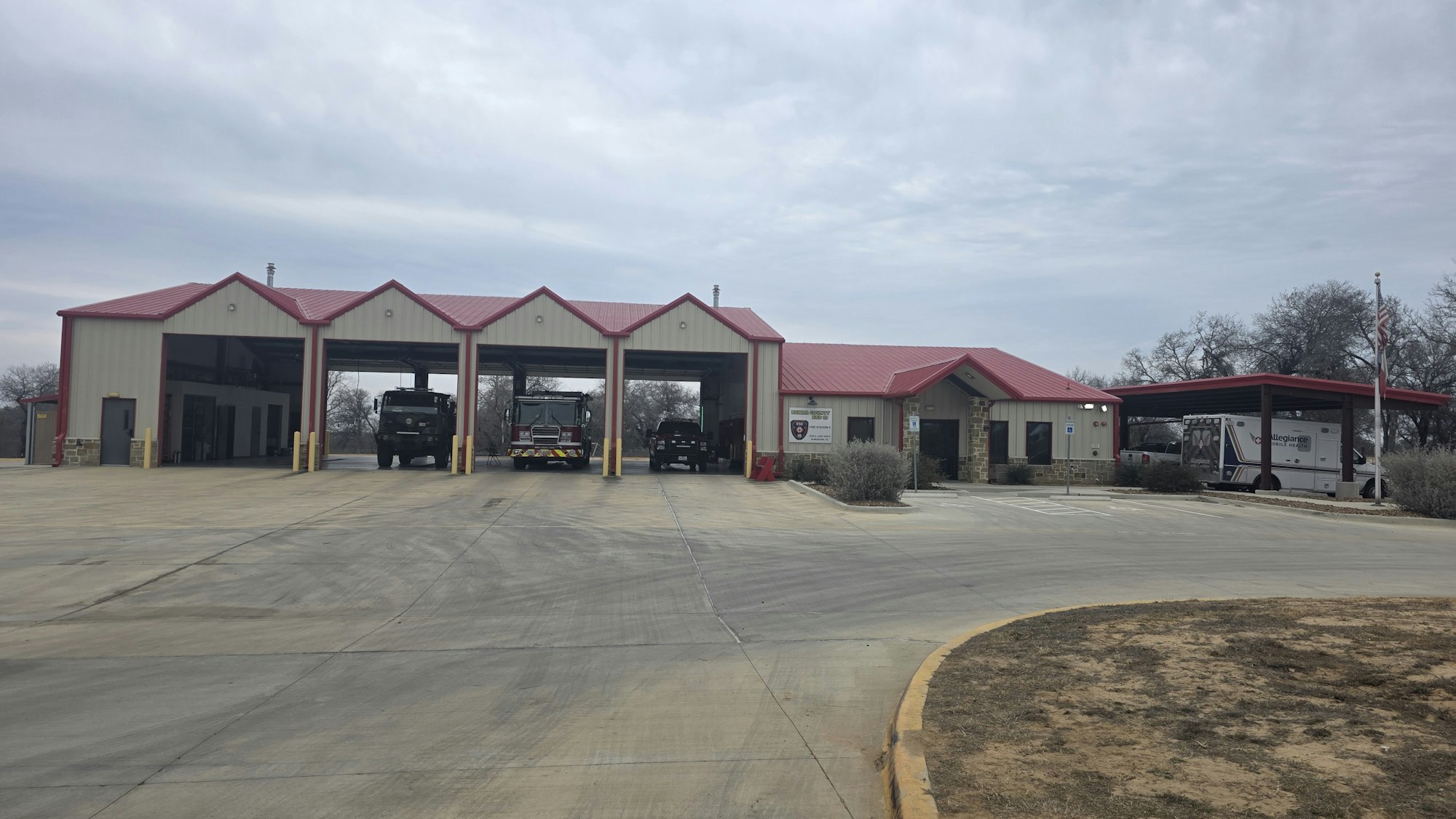The image shows a fire station with vehicles parked inside and outside, featuring a red roof and a modern design.