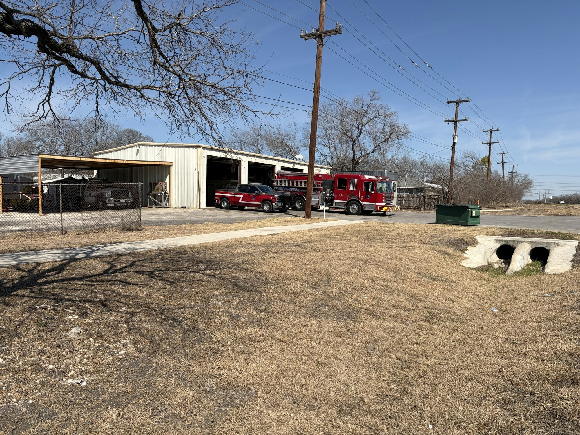A fire truck is parked outside a metal building, with power lines and a drainage area nearby, under a clear blue sky.