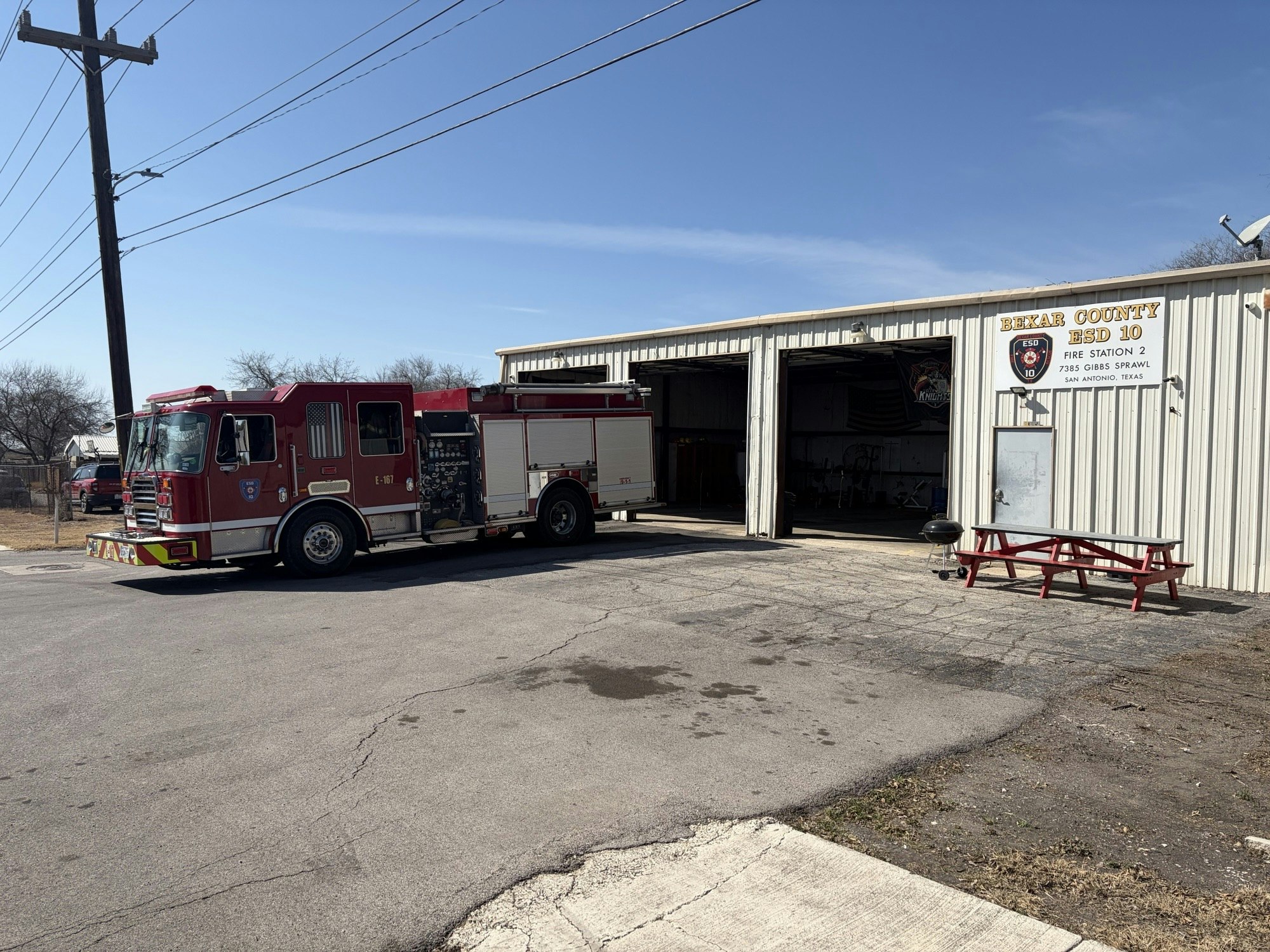 A red fire truck parked outside a fire station building with signage, picnic table, and clear blue sky in the background.
