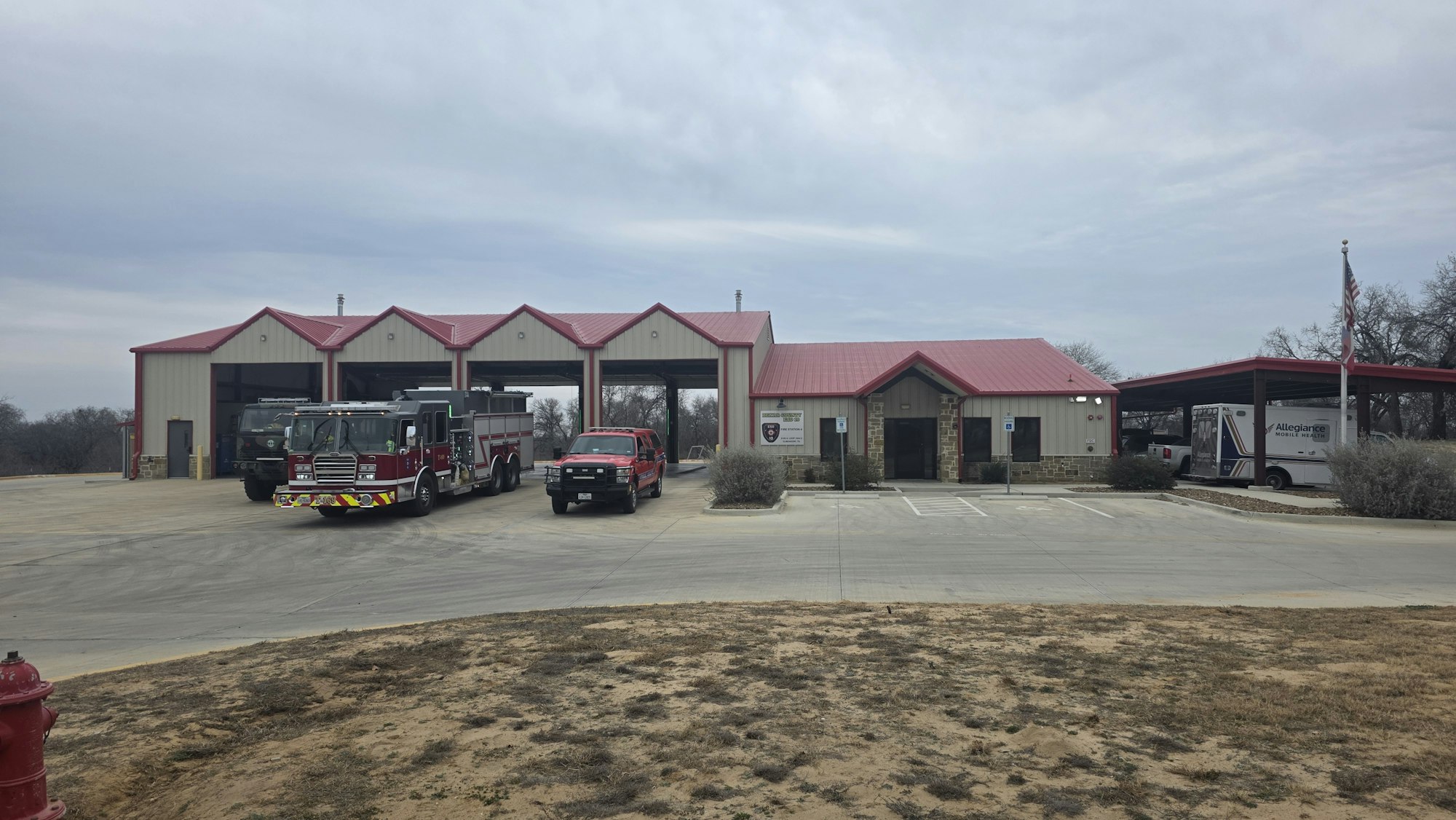 The image shows a fire station with fire trucks parked outside and an ambulance nearby, under a cloudy sky.