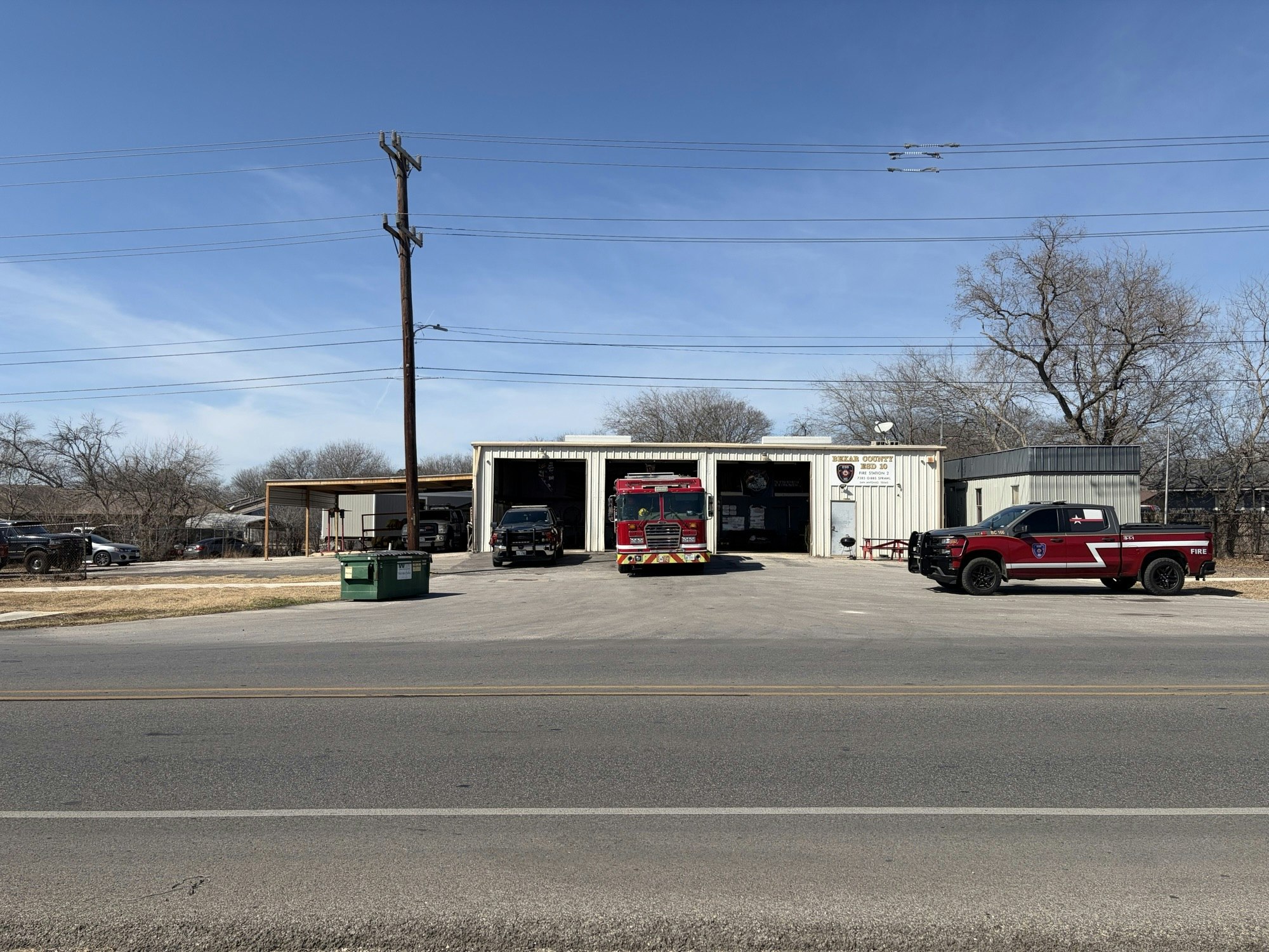A fire station with a fire truck and a pickup truck in front, surrounded by a parking area and a clear blue sky.