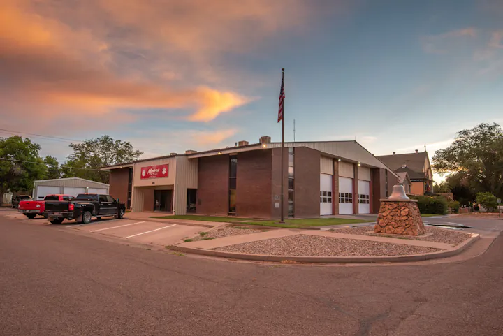 A building with a sign, parked trucks, a flagpole, a statue, and a colorful sky at dusk.