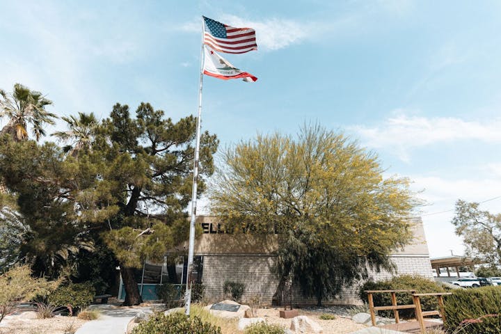 American and California flags flying above a building amidst trees on a sunny day.