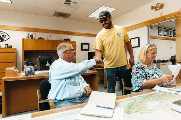Two men shake hands in an office while a woman reviews documents.
