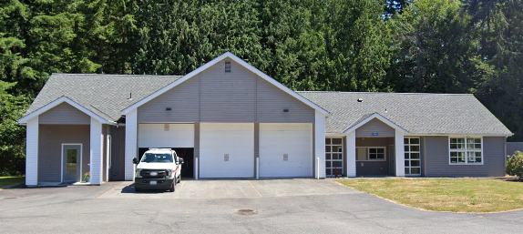 The image shows a medium-sized building with a gray exterior, large garage doors, and a white vehicle parked in front. Surrounded by trees.