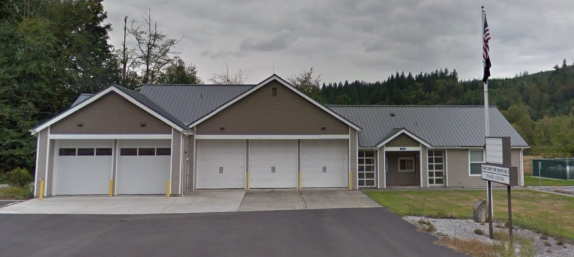 A building with a gray roof, garage doors, and a flagpole, surrounded by trees and greenery.