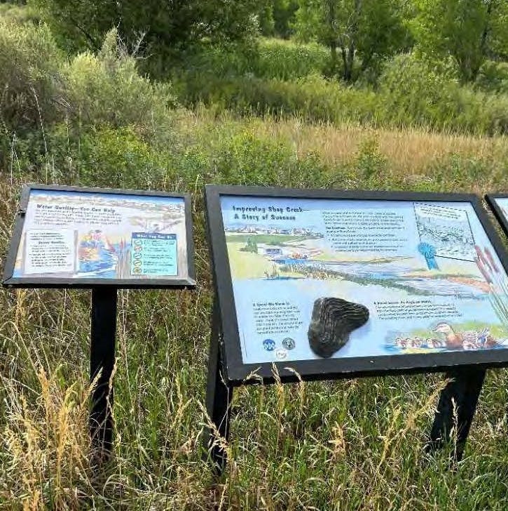 Outdoor informational signs about water quality and successful creek restoration, set in a grassy area with trees.