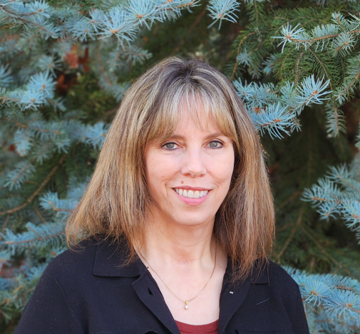 A person smiling in front of a background of green and blueish pine trees.