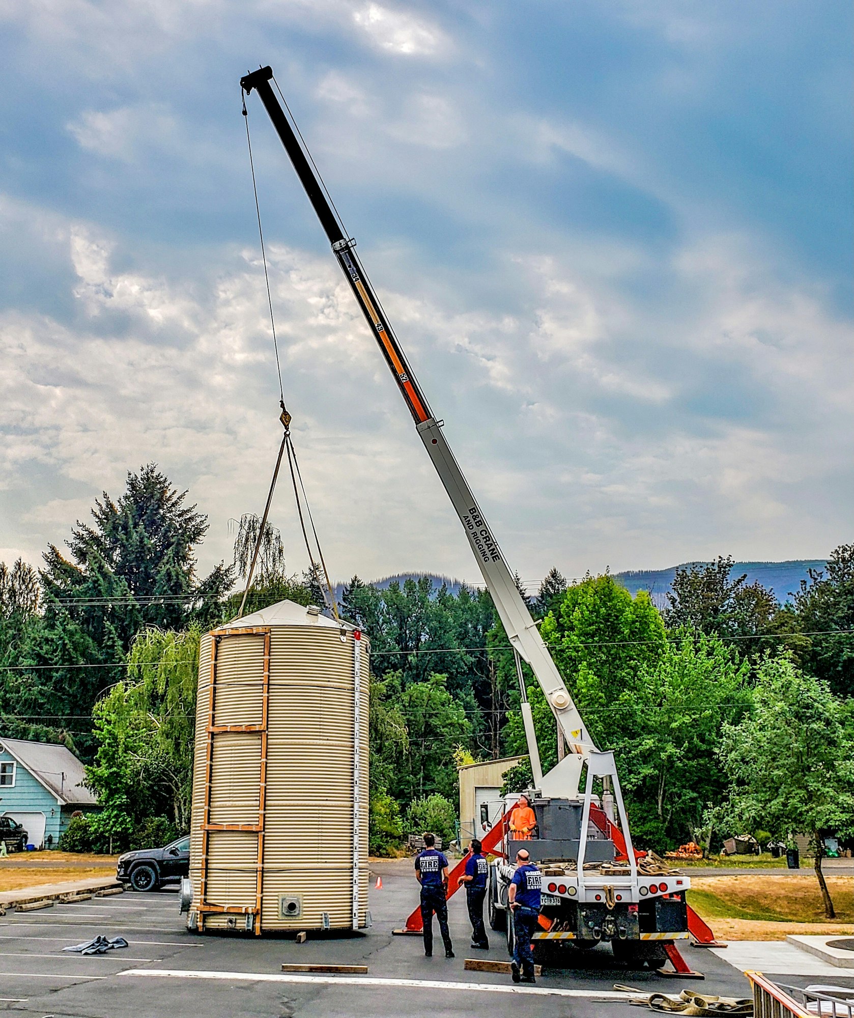 A crane is lifting a cylindrical structure while workers oversee the operation in a parking area with trees in the background.