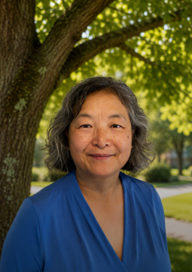 A smiling woman with gray hair wearing a blue top stands outdoors in front of a tree, surrounded by greenery.