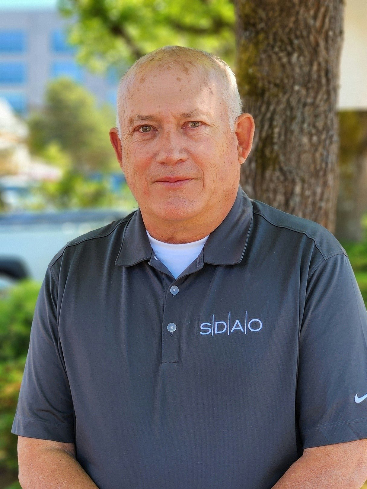 A man in a polo shirt with a logo, outdoor setting with trees and a building in the background.