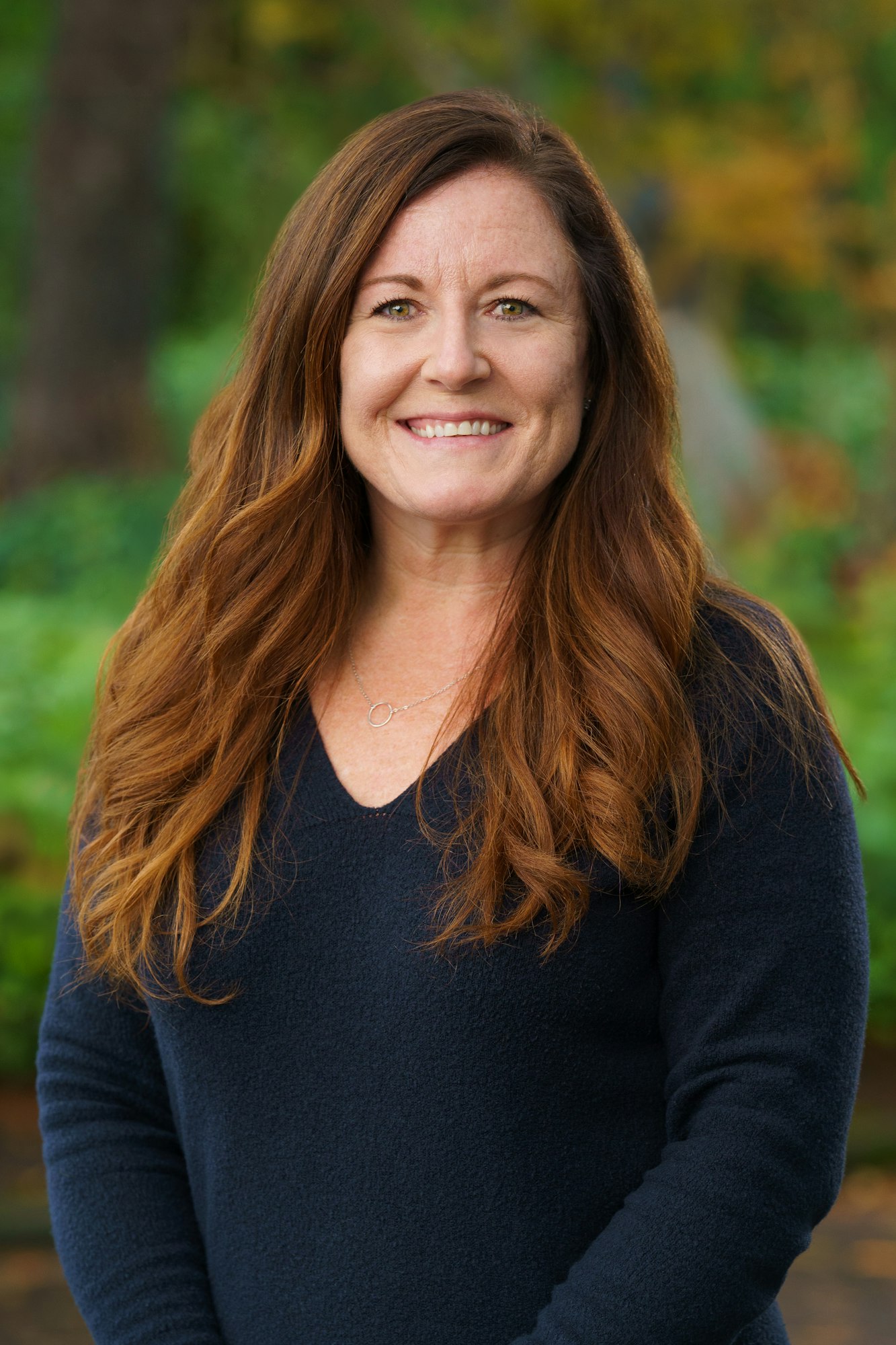 A woman with long, wavy hair is smiling, wearing a dark sweater, in a natural outdoor setting.