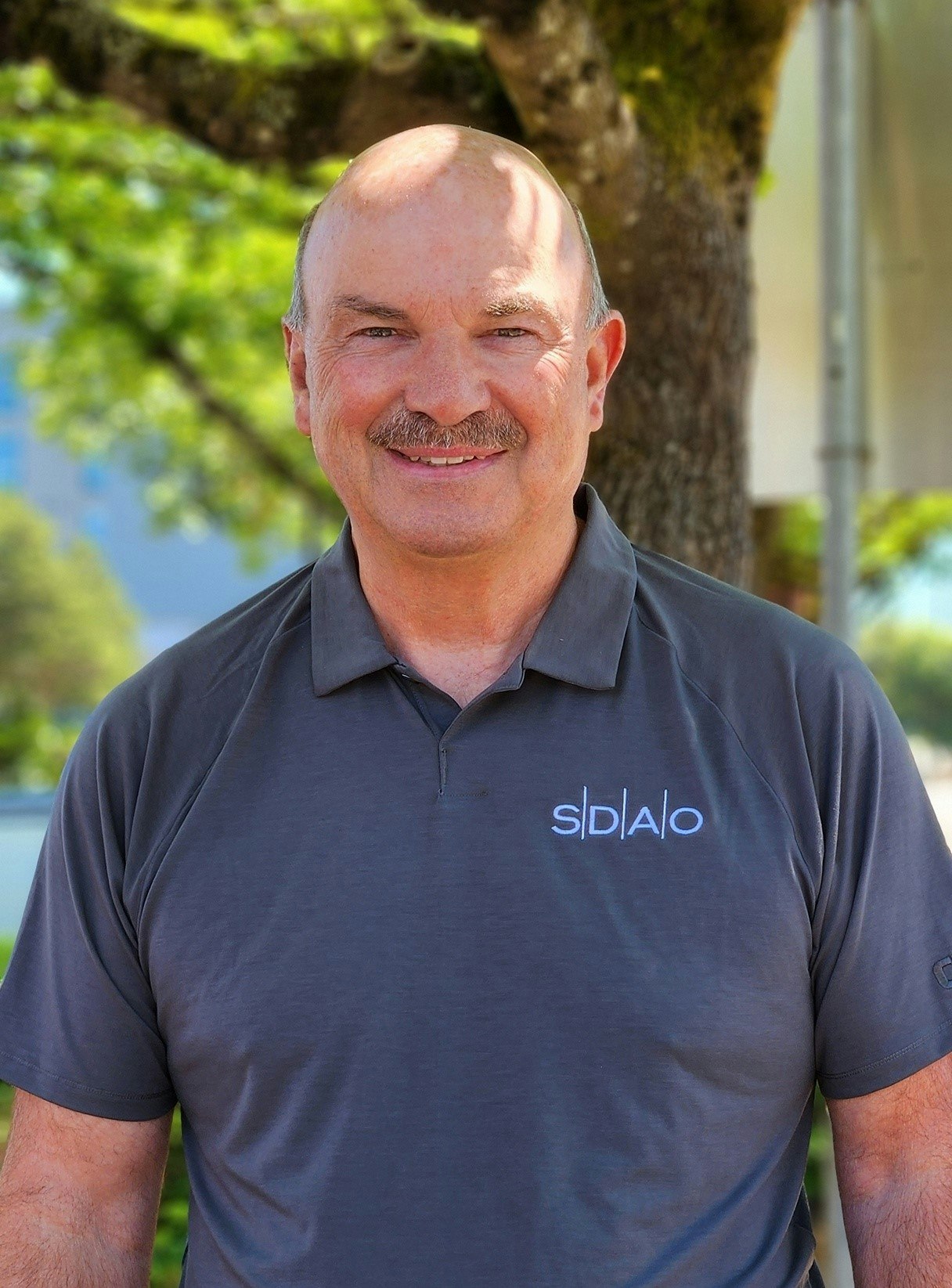A smiling man in a branded polo shirt standing outdoors with trees in the background.