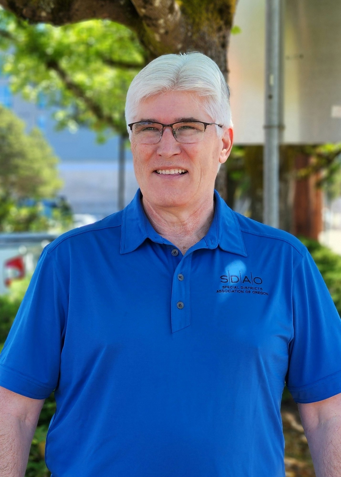 A smiling man in glasses wearing a blue polo shirt with a logo, standing outdoors.