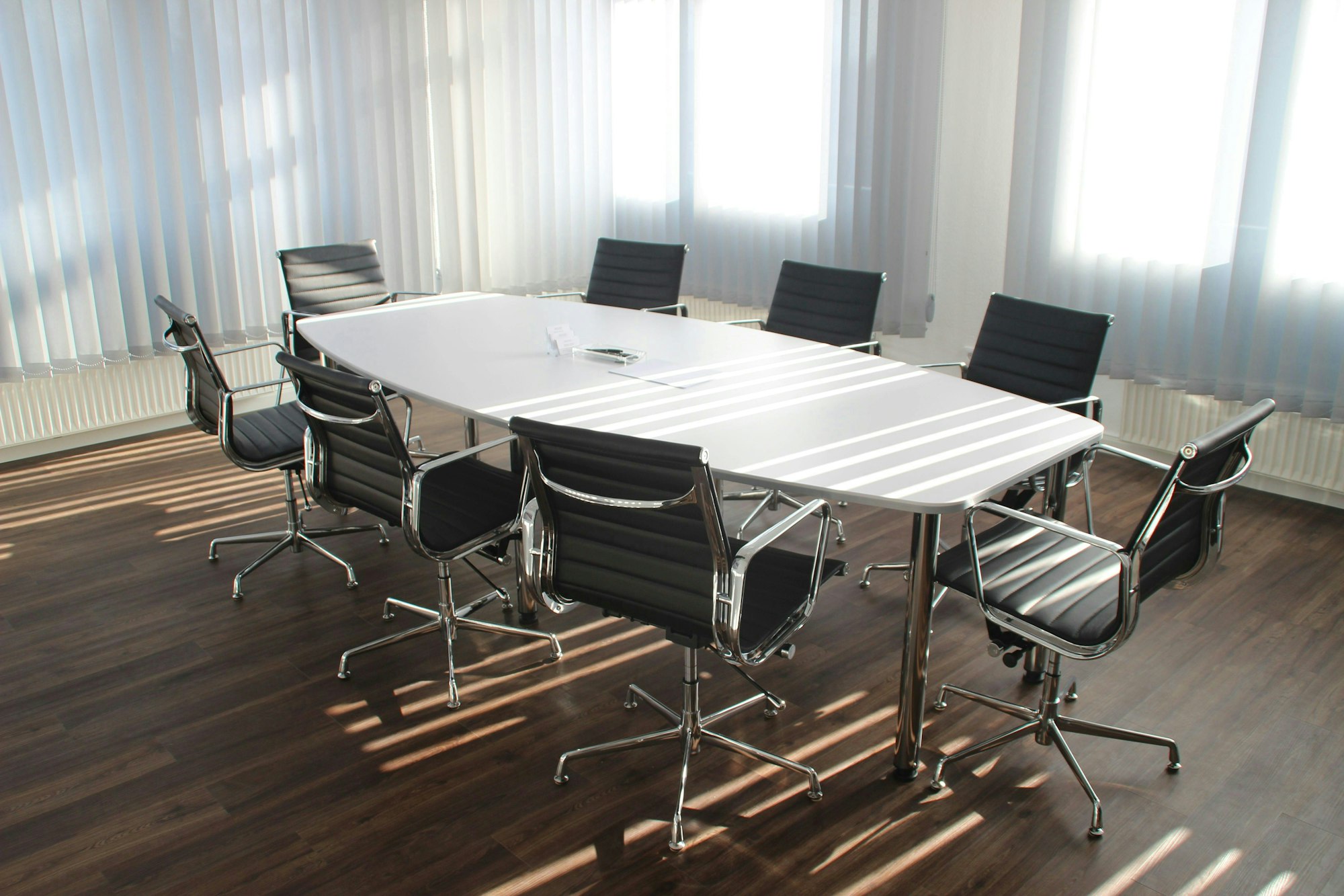 A modern conference room with a large white table and eight black chairs on a wooden floor, lit by natural light from windows.
