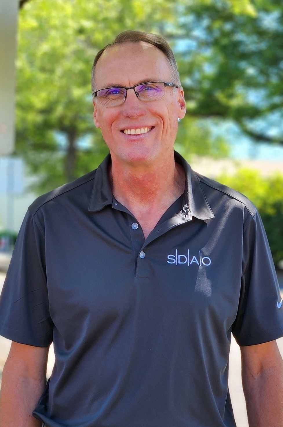 A smiling person in glasses wearing a polo shirt with a logo, standing before a blurred green background.