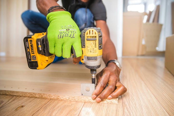 Person using a yellow cordless drill to secure a piece to wooden flooring.