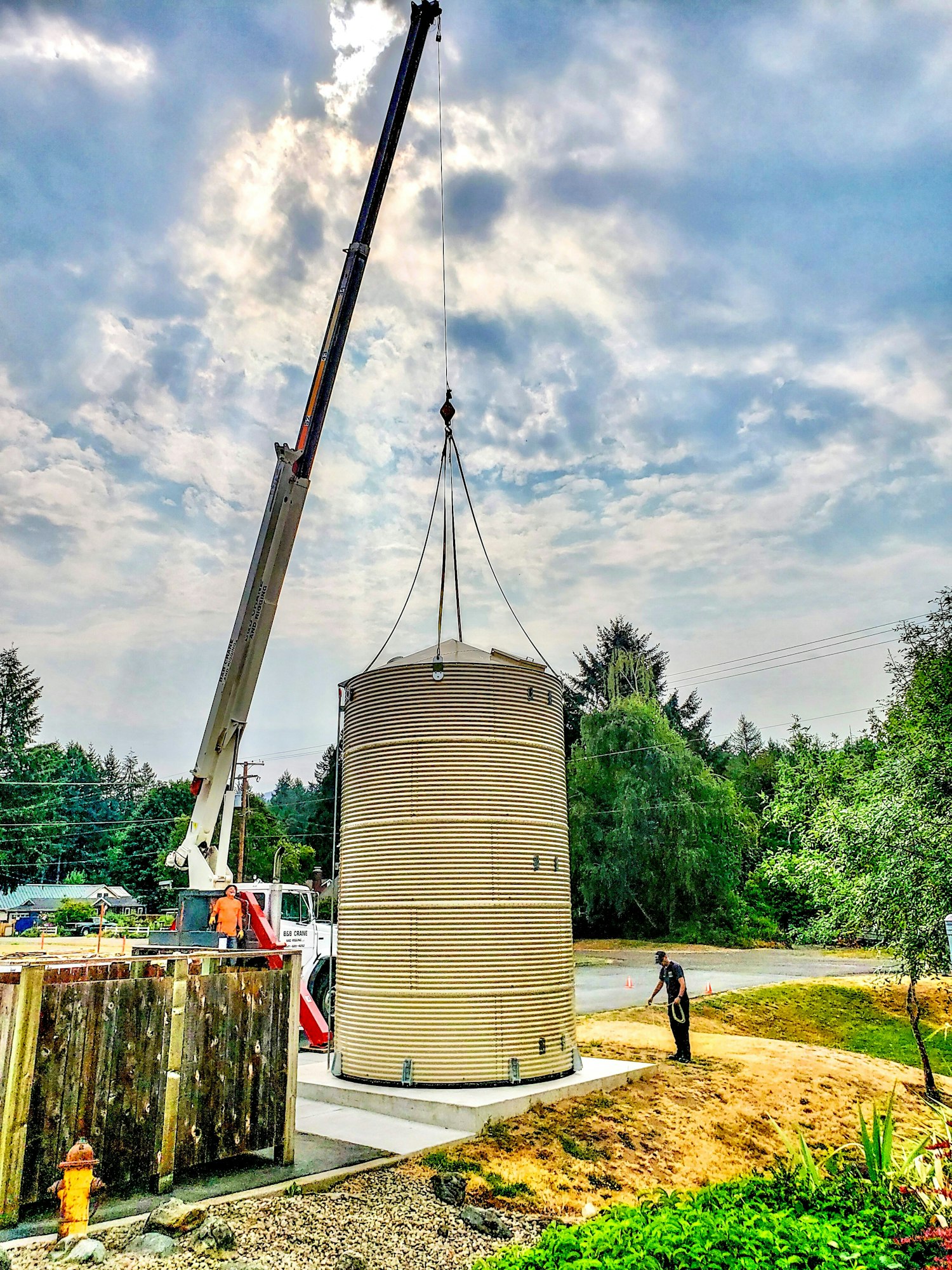 A crane lifts a cylindrical structure outdoors, with workers nearby and a cloudy sky above.