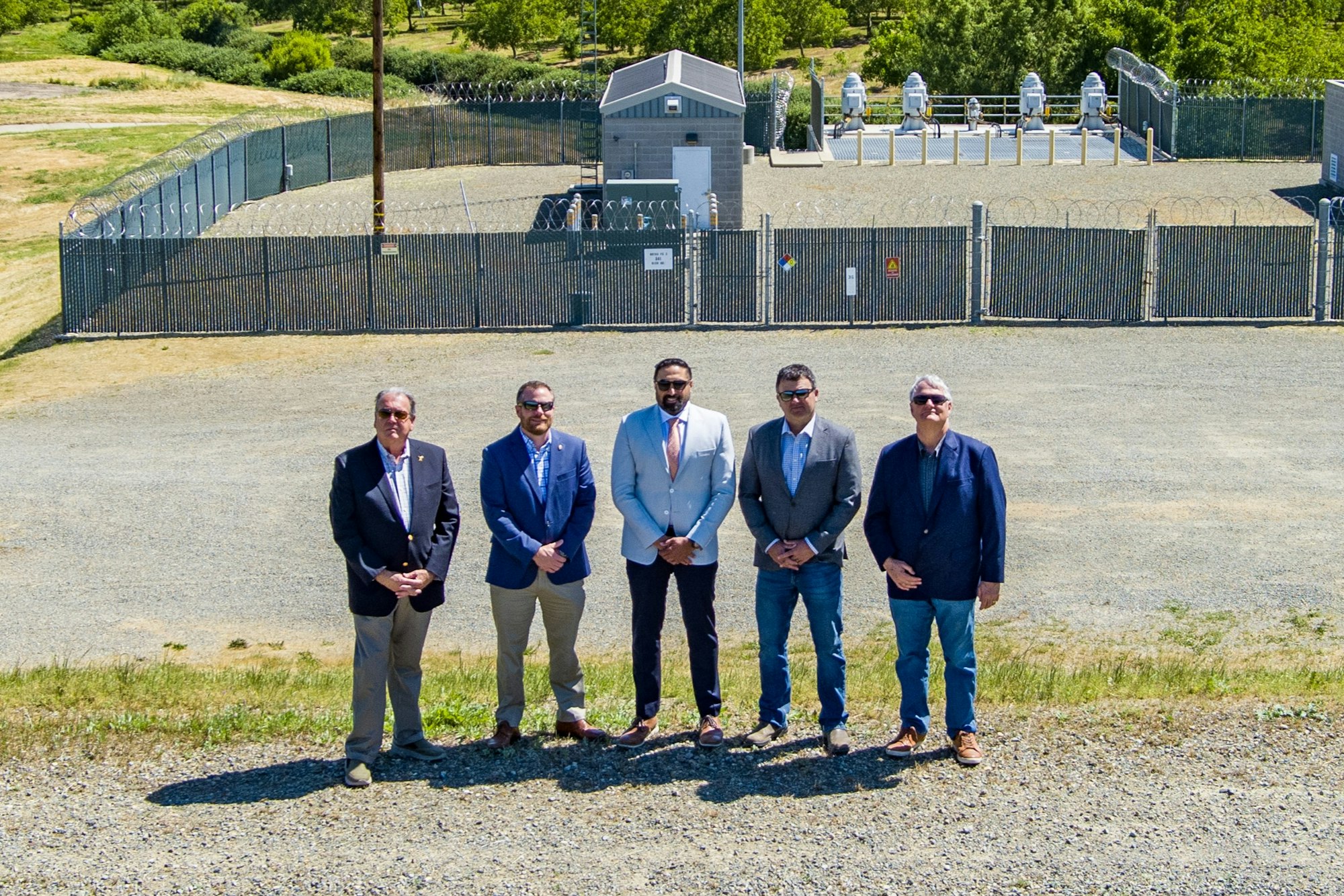 Five people in suits stand in front of a fenced industrial facility surrounded by greenery.