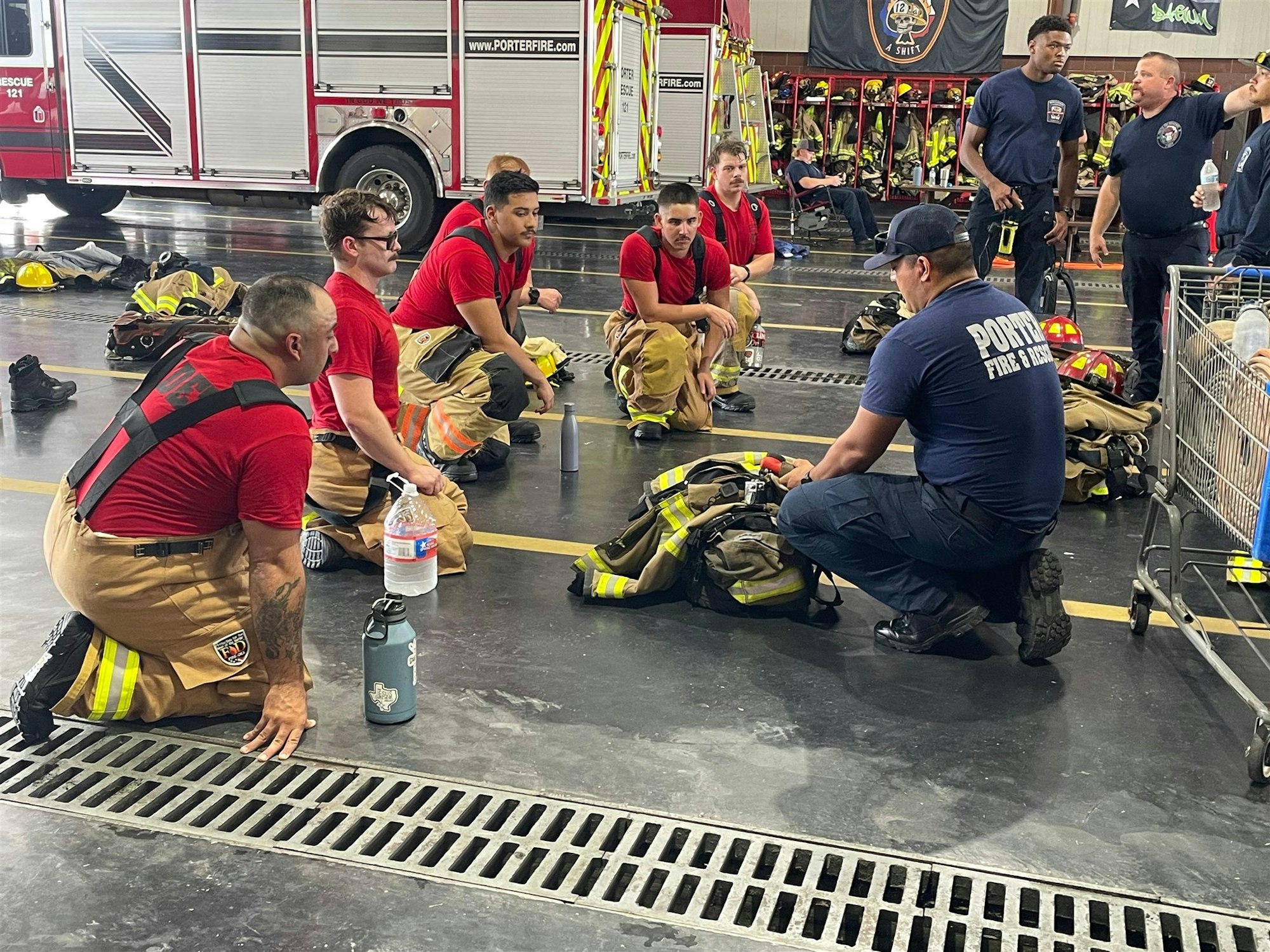 A group of firefighters in training is seated, listening to an instructor in a fire station setting.
