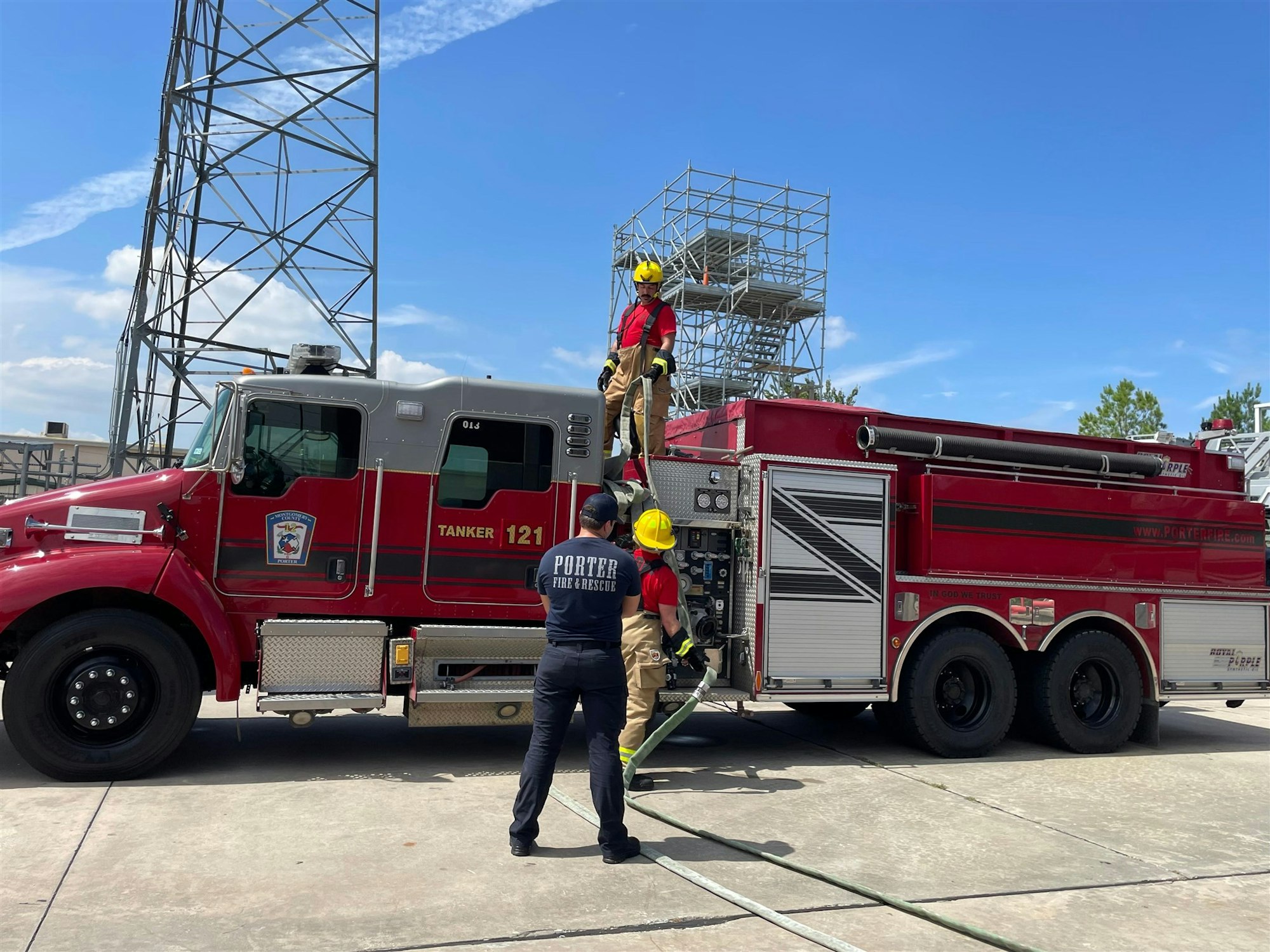 Firefighters practicing on a red tanker truck, with one climbing the vehicle and another managing hoses in bright weather.
