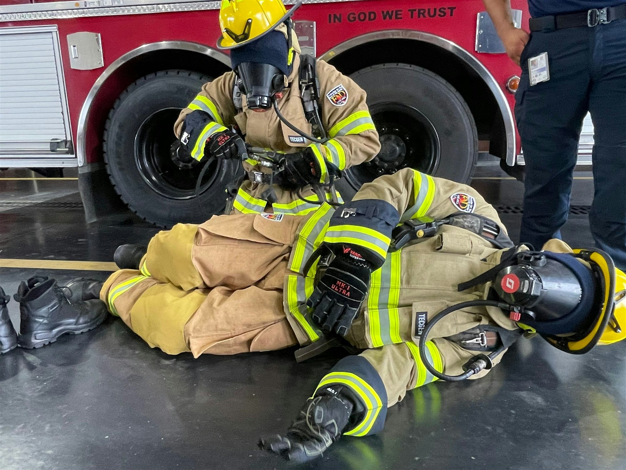 Two firefighters in full gear practice a rescue operation near a fire truck. One is providing assistance to another.