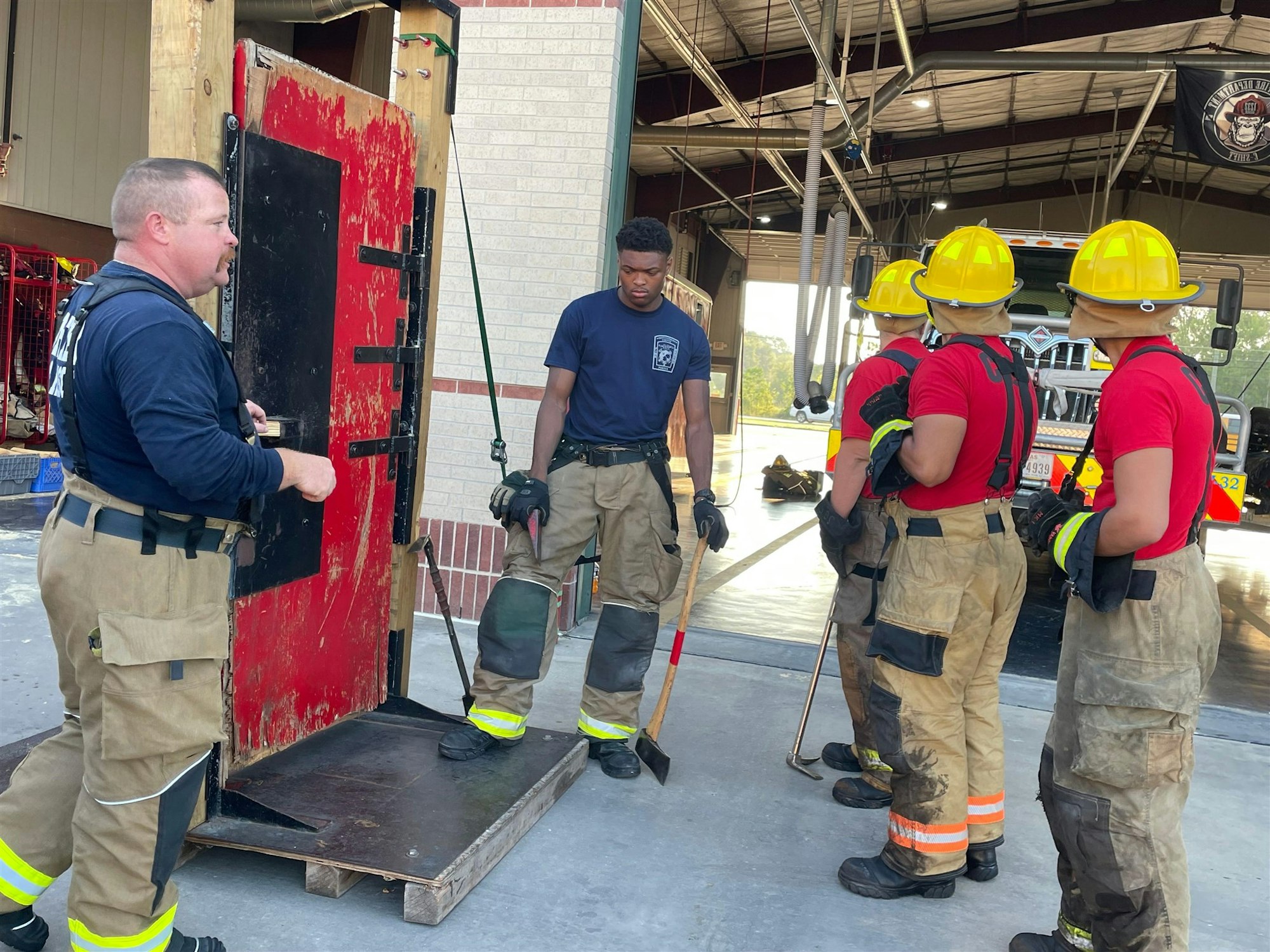 A group of firefighters in training stands near a red practice door, learning tools and techniques for emergency situations.