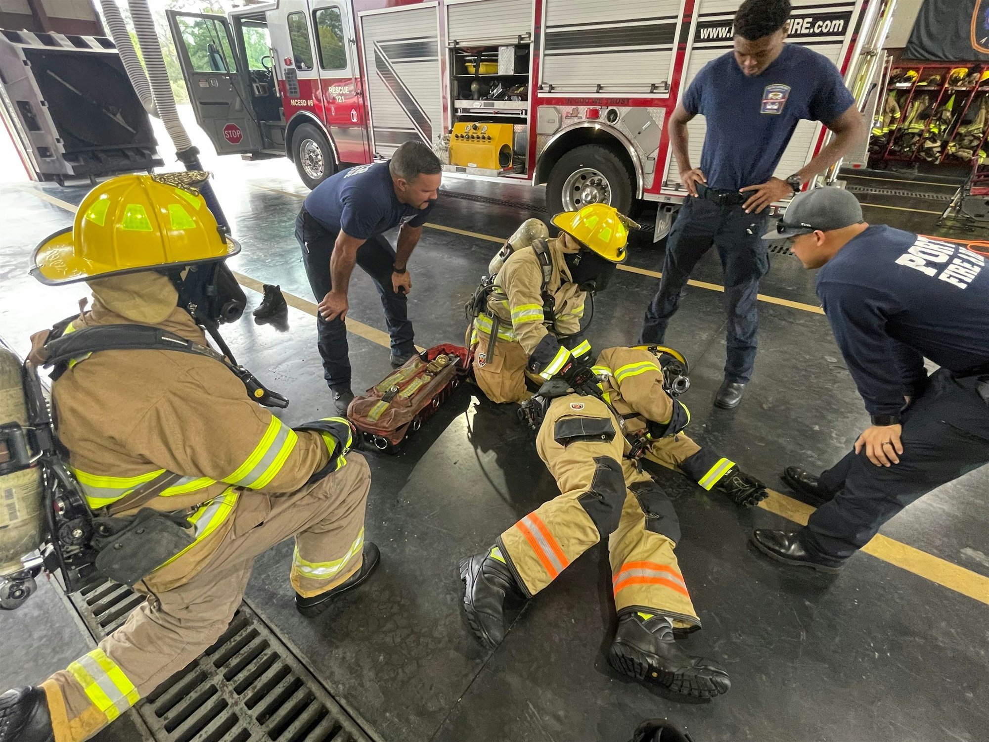Firefighters are training or assisting each other in a fire station, with one individual on the ground receiving help.