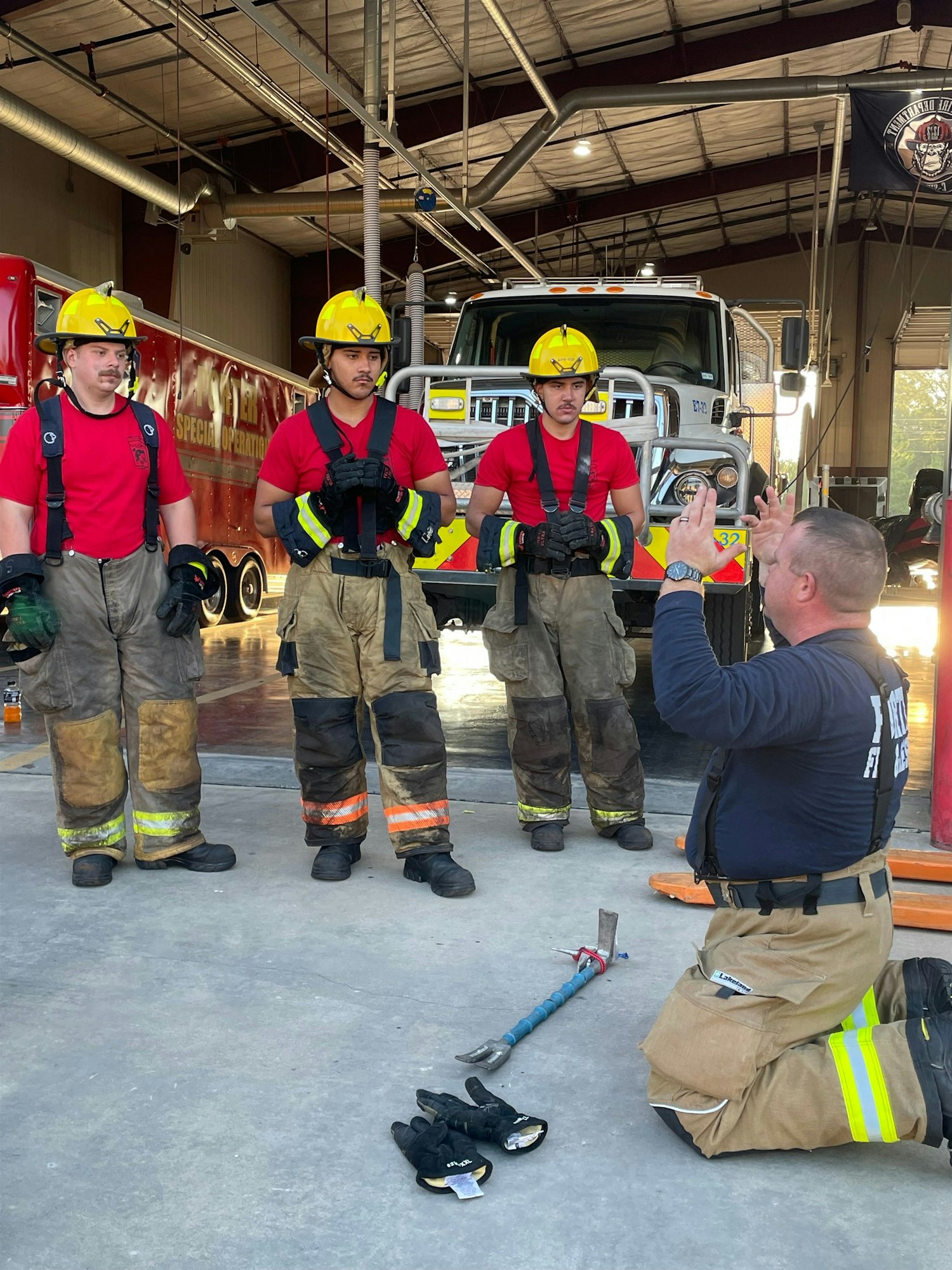 A group of firefighters in training listens to an instructor in a fire station, with gear and tools laid out on the floor.