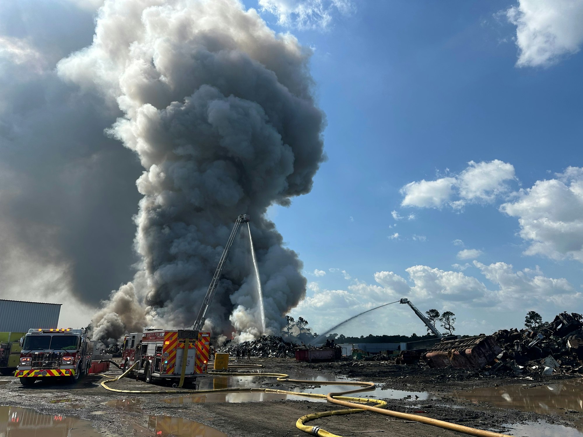 Firefighters battle a large industrial blaze with thick smoke under a blue sky.