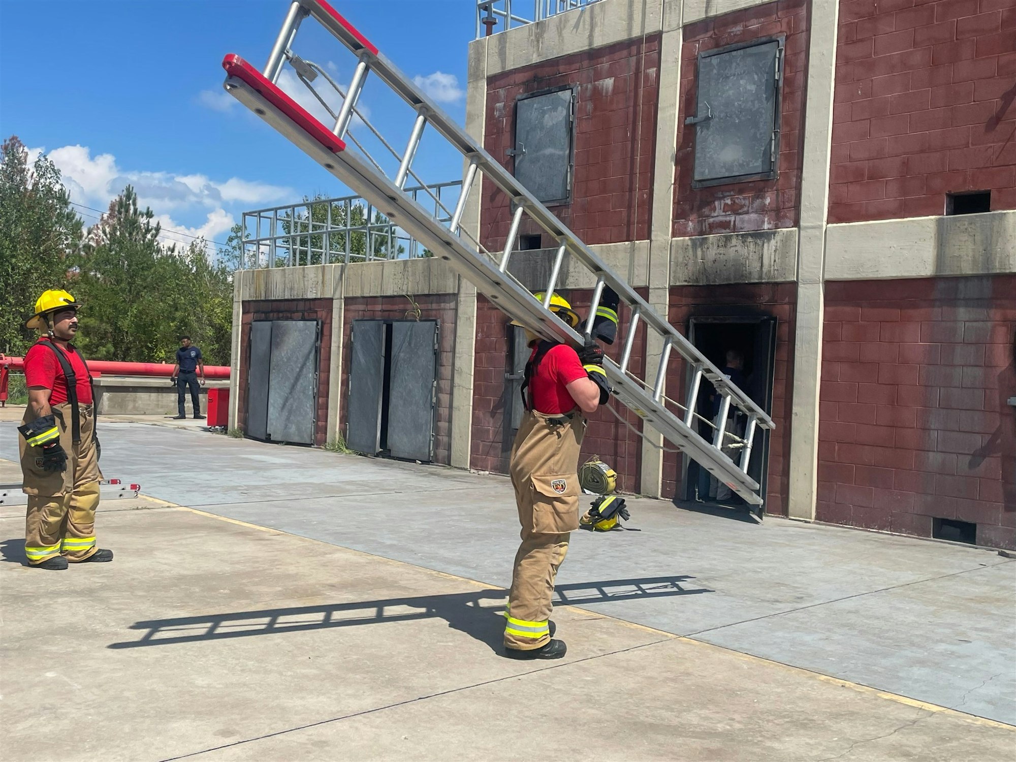 A training scenario for firefighters, featuring two in gear, one carrying a ladder, near a building with smoke stains.
