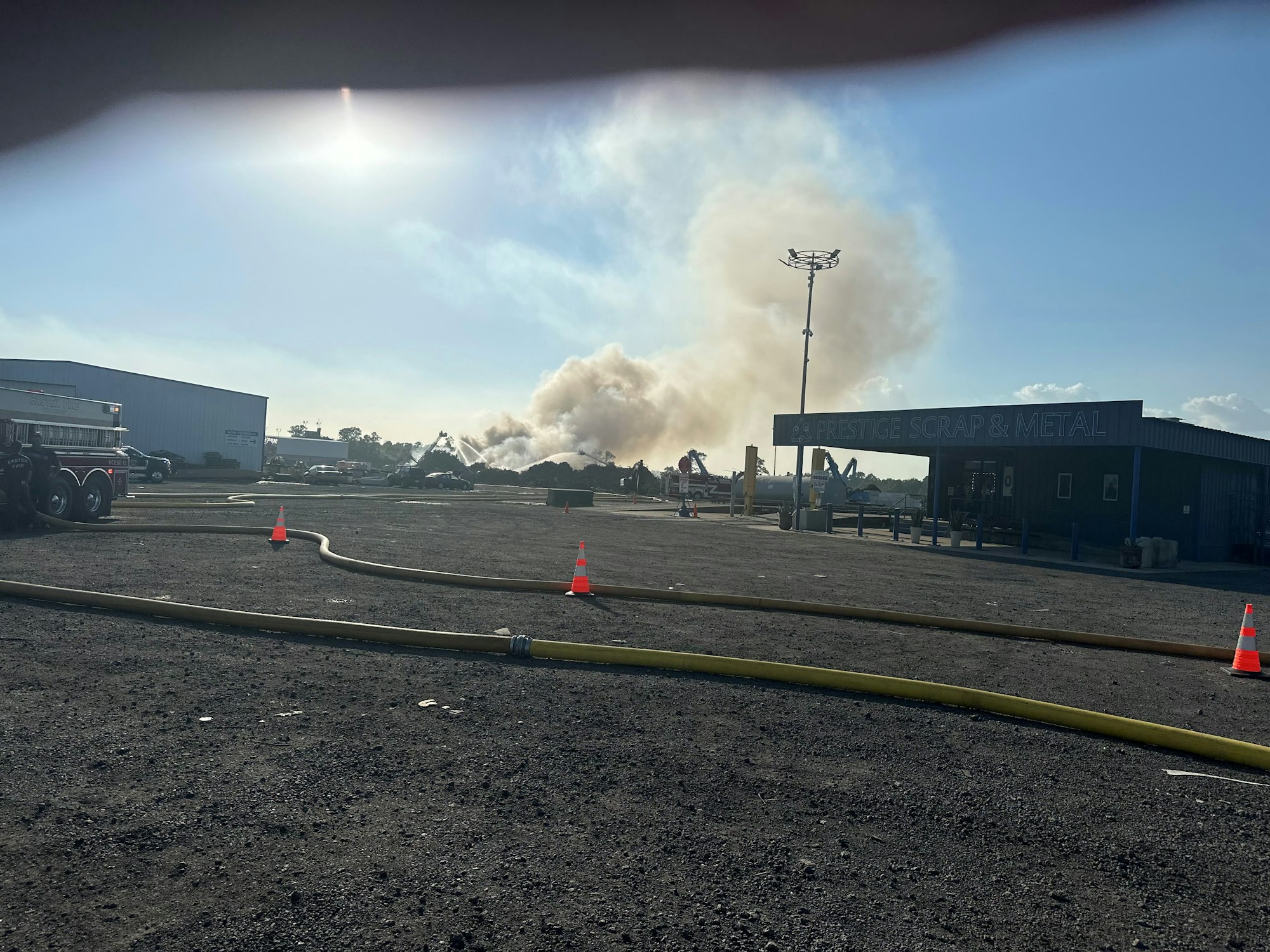 A fire at Prestige Scrap & Metal, with smoke, fire hoses on the ground, and emergency response underway.