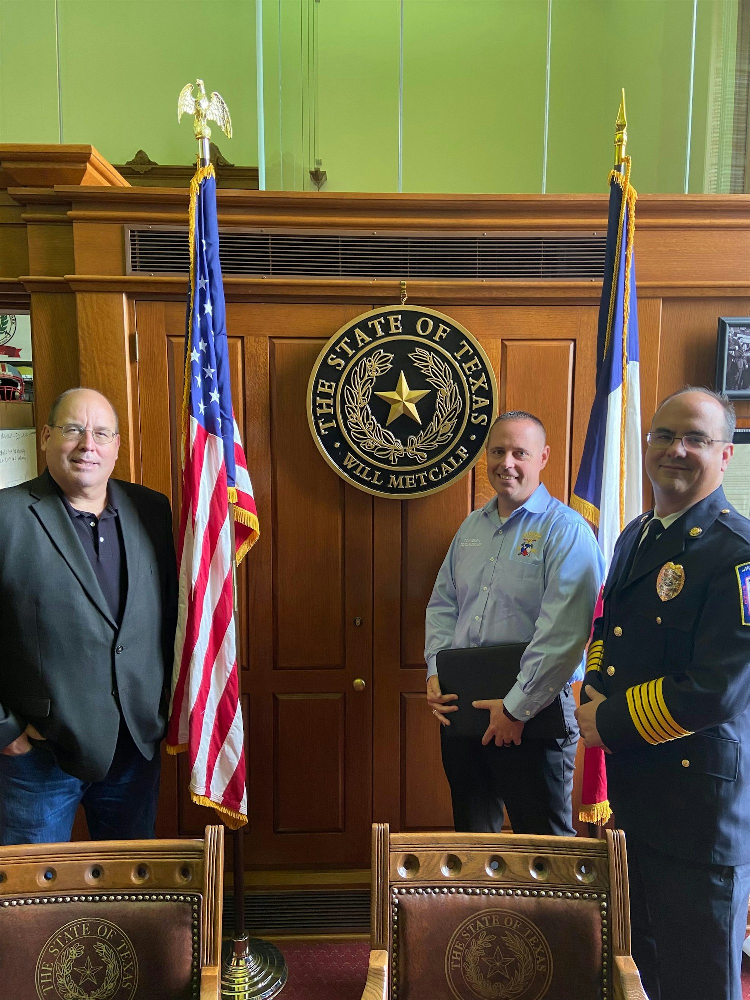 Three men stand in a Texas government office by the state seal and flags, with wooden furniture visible in the background.