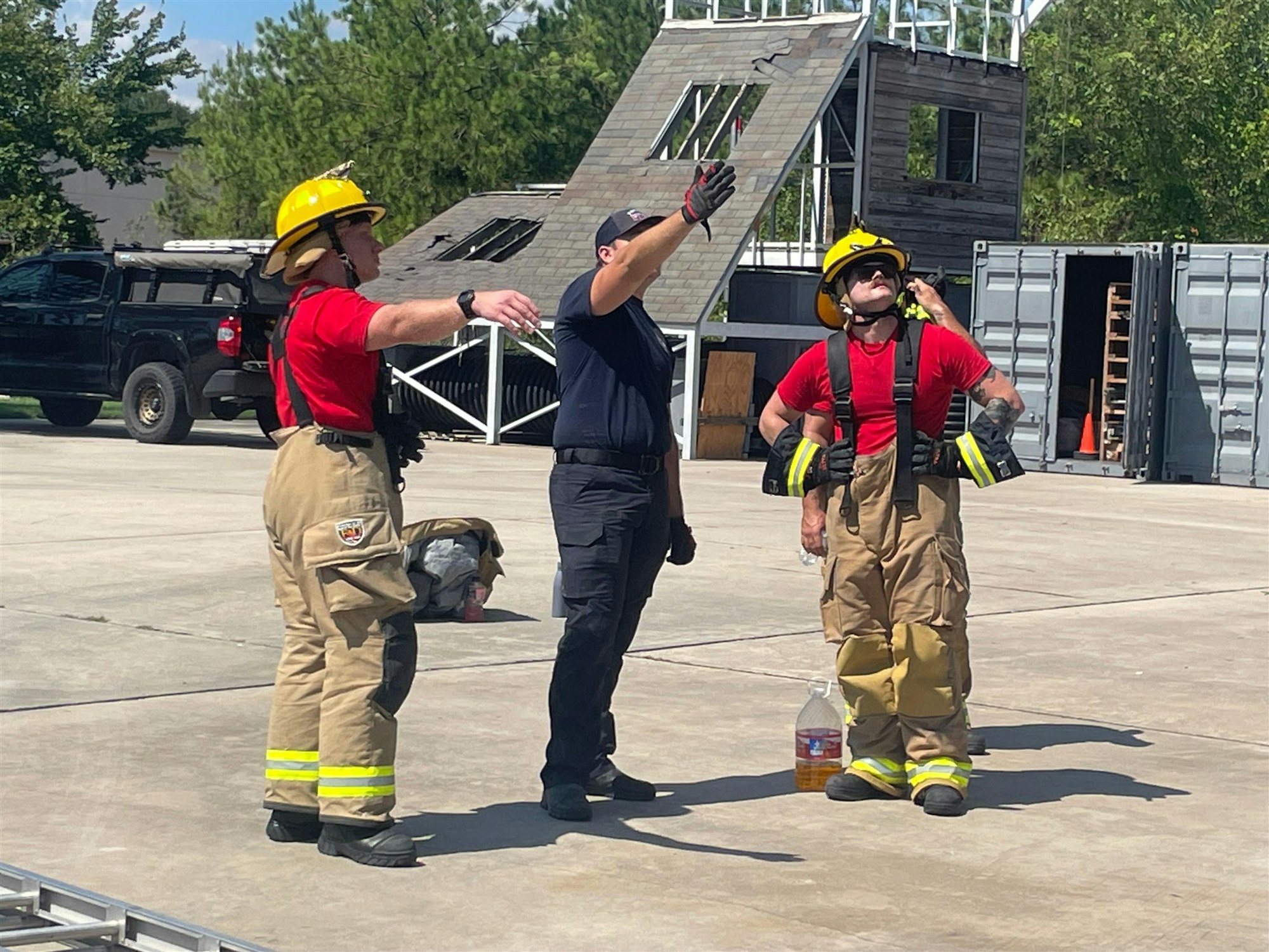 Three firefighters in gear discuss training or a drill in an outdoor setting, with training structures and equipment visible.