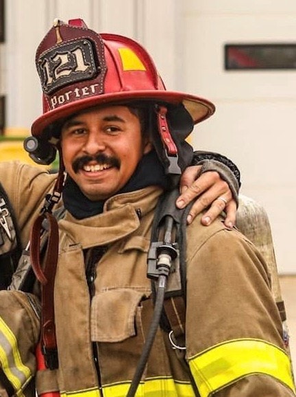 A smiling firefighter in full gear, wearing a helmet with the number 121, stands with a hand on his shoulder.