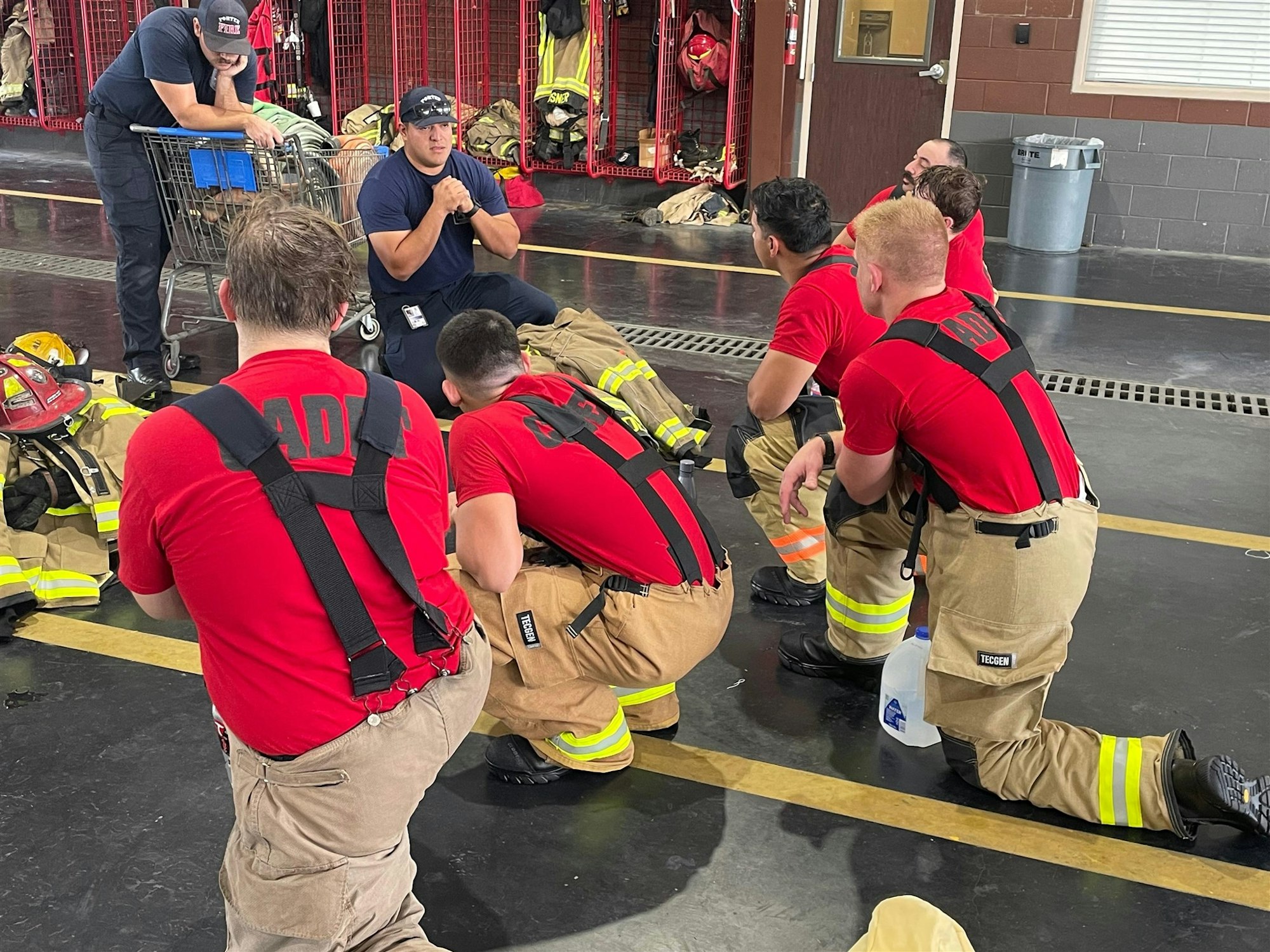A group of firefighters in training listens to an instructor at a fire station, surrounded by gear and equipment.
