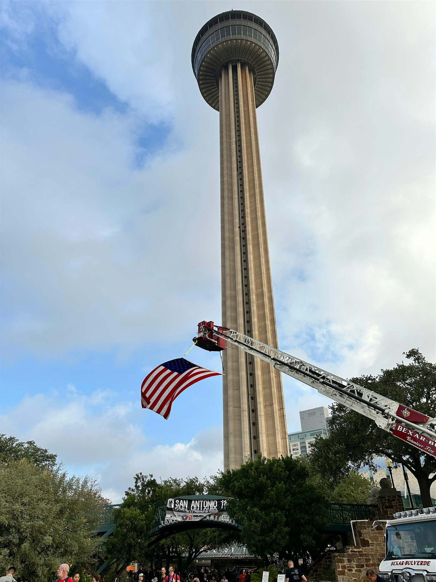 A tall tower, possibly in San Antonio, with a fire truck raising an American flag, and a crowd below.