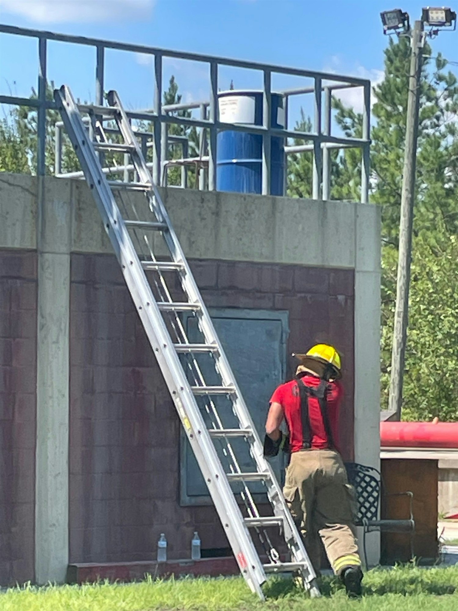 A firefighter in a red shirt and yellow helmet is climbing a ladder next to a building, with bottles of water visible nearby.