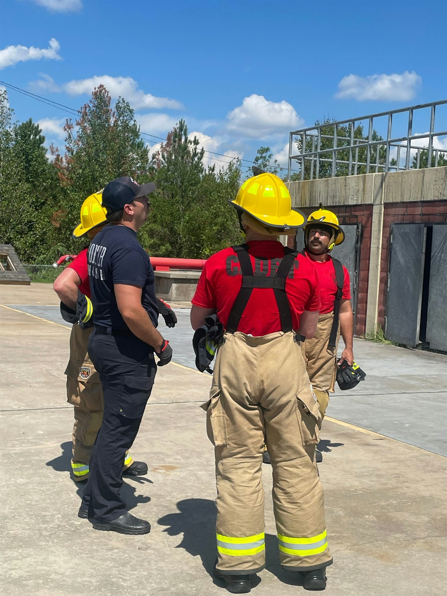 Four individuals, some in firefighting gear, gather outdoors under a blue sky, possibly discussing training or safety procedures.