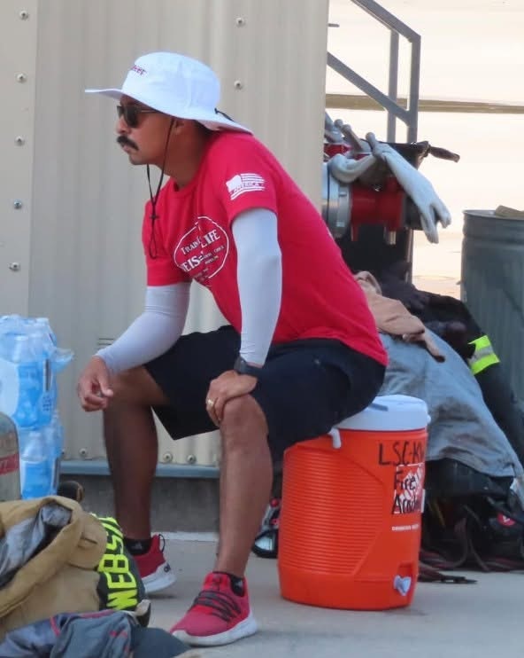 A man in a red shirt and bucket hat sits on a cooler, resting among gear and equipment.
