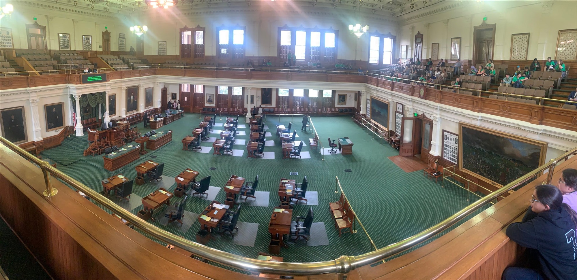 The image shows a spacious legislative chamber with wooden furniture, green carpeting, and an audience in an upper gallery.