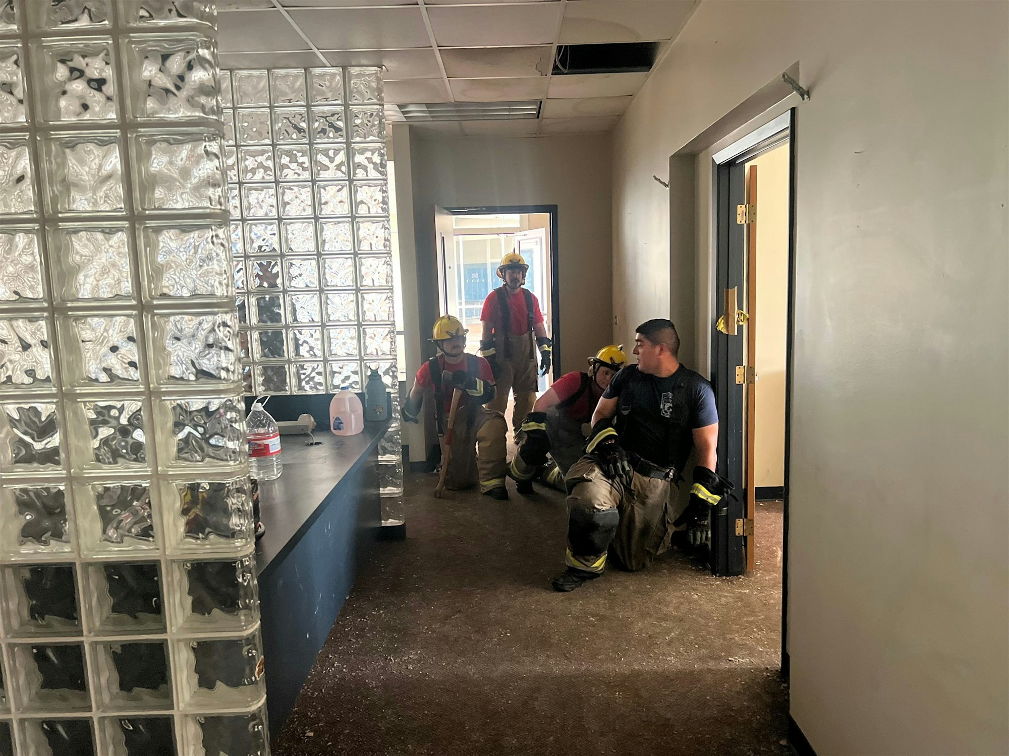 Firefighters rest in a hallway featuring glass block walls, with a table holding a water jug in a partially empty building.