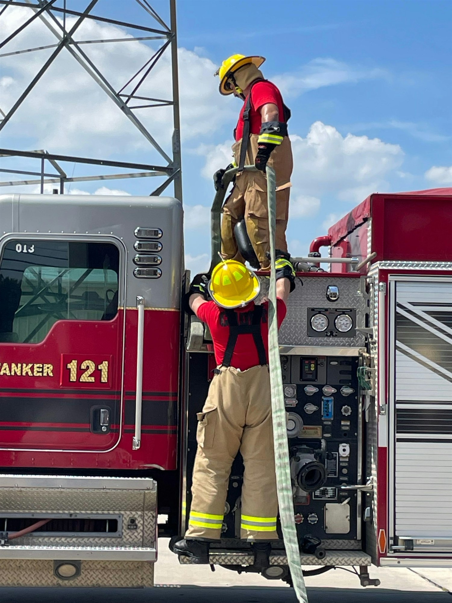 Two firefighters in uniforms work on a fire truck, handling equipment under a clear blue sky.