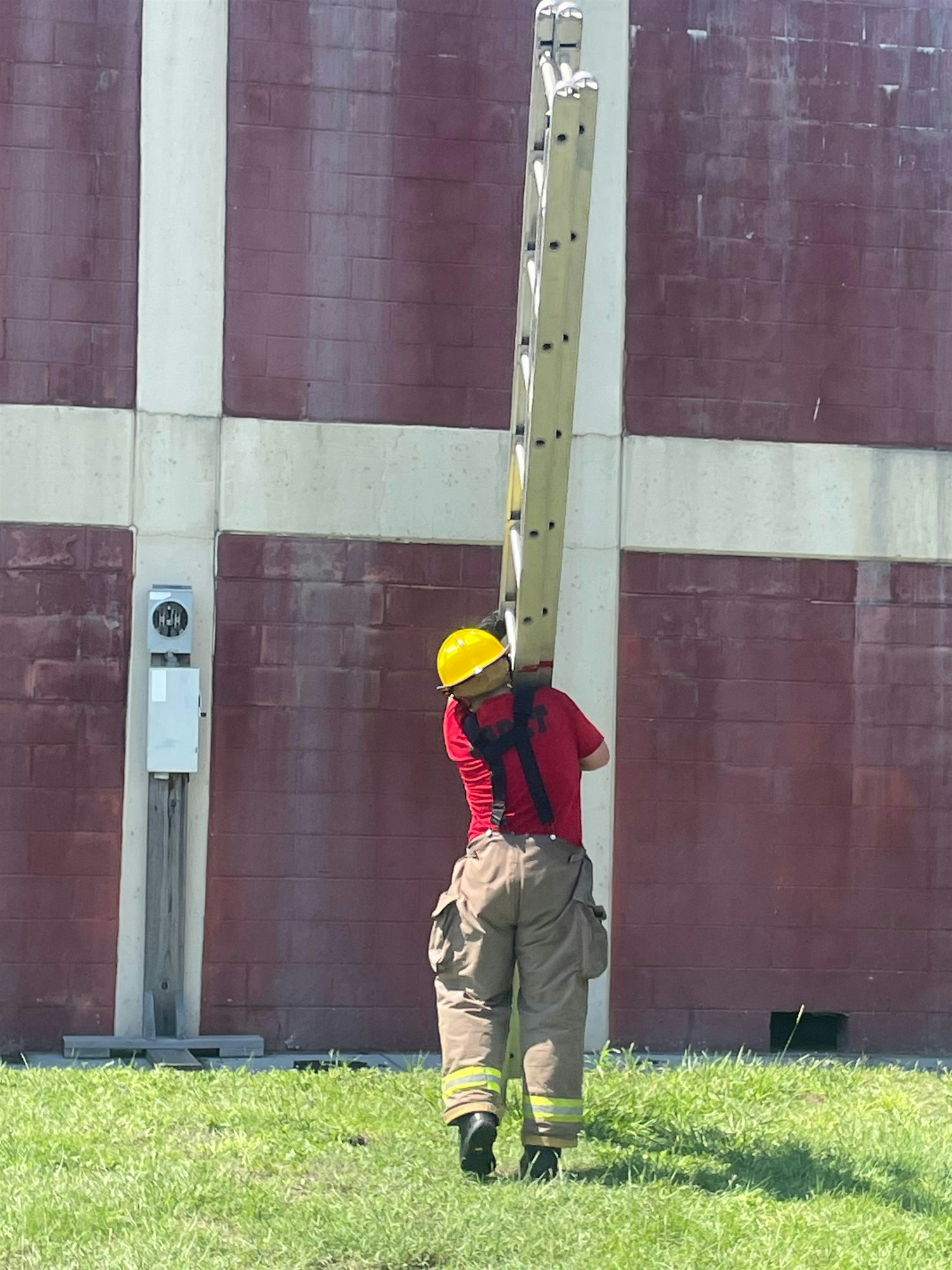 A person in firefighting gear carries a ladder across a grassy area near a red building.