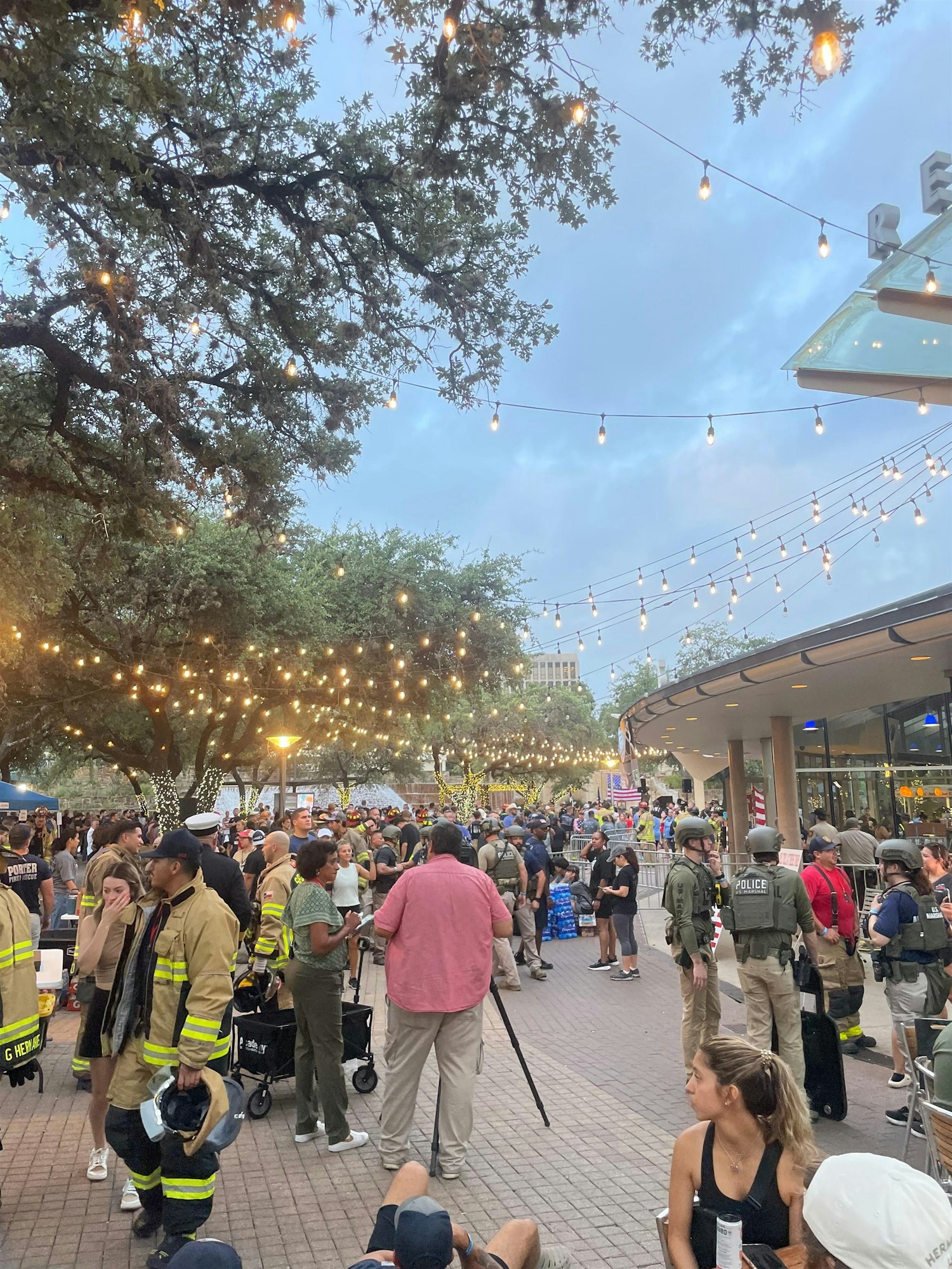 A lively outdoor scene with crowds, string lights, and a mix of uniformed personnel, suggesting a community event or gathering.