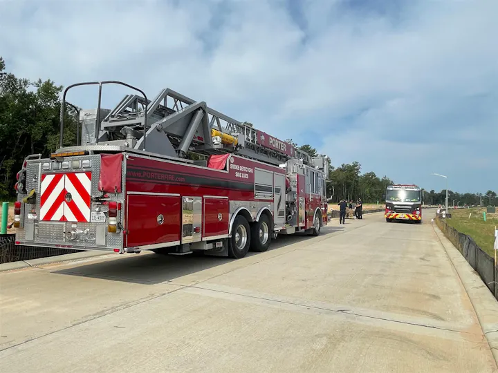 The image shows two fire trucks parked on a road, with firefighters present and a construction area in the background.