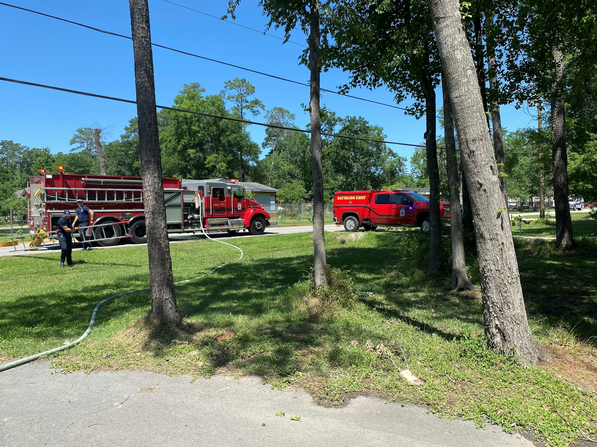 A fire truck, a "Battalion Chief" vehicle, firefighters, trees, and hoses on a sunny day.